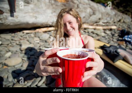 Junge Frau sitzt am felsigen Strand, hält Getränke, lächelnd, Short Sands Beach, Oregon, USA