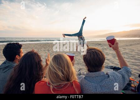 Gruppe von Freunden sitzen am Strand, gerade Freund tun Wagenräder auf und Sonnenuntergang, Rückansicht Stockfoto