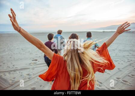 Gruppe von Freunden zu Fuß am Strand, junge Frau mit den Armen in der Luft, Rückansicht Stockfoto