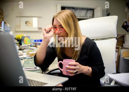 Junge Frau im Büro, am Schreibtisch, mit Kaffeetasse, Brille Stockfoto