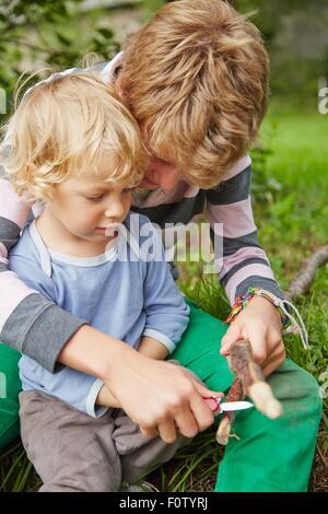 Junge sitzt mit jüngeren Bruder mit einem Taschenmesser auf Zweig im Garten Stockfoto