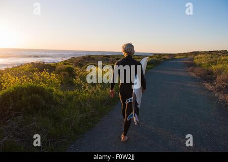 Junge männliche Surfer bei Sonnenaufgang mit Surfbrett auf Landstraße an Küste Stockfoto