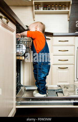 Niedliche Baby stehend auf Geschirrspüler in der Küche und hilft es entladen Stockfoto