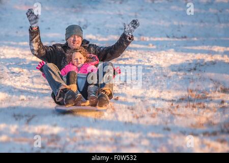 Porträt des Lächelns, Vater und Tochter fahren Schlitten bergab im Schnee Stockfoto