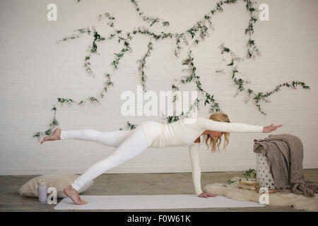 Eine blonde Frau auf einer weißen Matte in einem Raum, ihr Bein und Arm ausgestreckt. Eine Schlingpflanze Pflanze an der Wand hinter ihr. Stockfoto