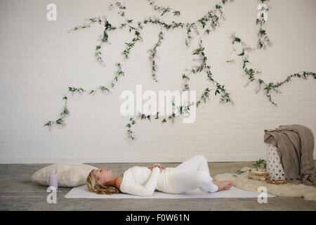 Eine blonde Frau in einem weißen Trikot und Leggings, liegend auf einer weißen Matte in einem Raum. Eine Schlingpflanze Pflanze an der Wand hinter ihr. Stockfoto