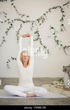 Eine blonde Frau sitzt auf einer weißen Matte in einem Raum, streckte ihre Arme. Eine Schlingpflanze Pflanze an der Wand hinter ihr. Stockfoto