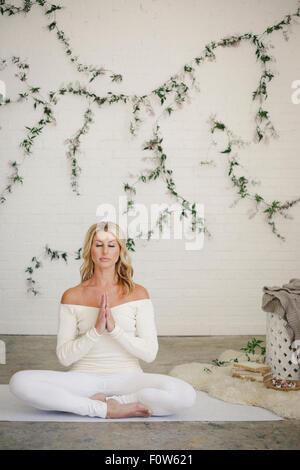 Eine blonde Frau sitzt auf einer weißen Matte in einem Raum, Yoga zu tun. Eine Schlingpflanze Pflanze an der Wand hinter ihr. Stockfoto