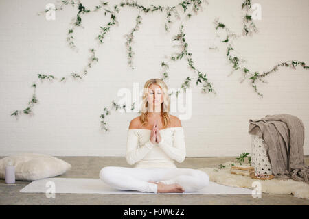 Eine blonde Frau sitzt auf einer weißen Matte in einem Raum, Yoga zu tun. Eine Schlingpflanze Pflanze an der Wand hinter ihr. Stockfoto