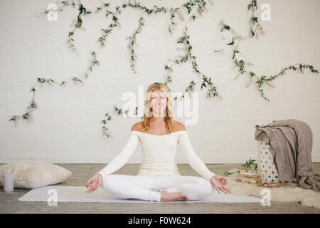 Eine blonde Frau in einen weißen Turnanzug sitzt auf einer weißen Matte in einem Raum, Yoga zu tun. Eine Schlingpflanze Pflanze an der Wand hinter ihr. Stockfoto