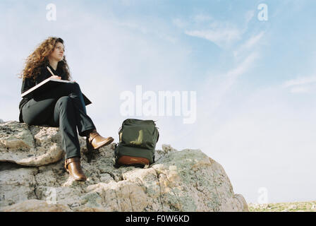 Eine Frau sitzt auf einem Felsen, schreiben in einem Notebook, einem Rucksack liegen durch ihre Füße. Stockfoto