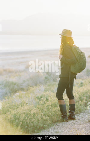 Eine Frau trägt einen Hut und Rucksack, stehend auf einem Weg mit Blick auf einen See und Tal.