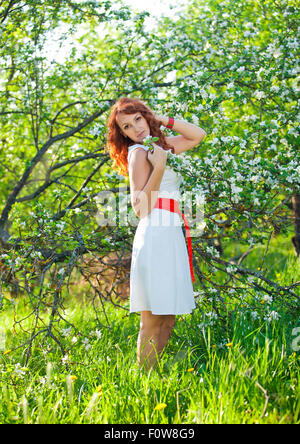 Freie glückliche Frau mit wunderschönen roten Haaren, die Natur genießen. Schönheit Mädchen im Freien im Frühlingsgarten. Freiheit-Konzept. Gesundes lächelndes Mädchen über grüne Blumen Natur Hintergrund. Apfelbäume in Blüte Stockfoto
