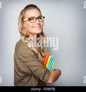 Porträt von Süße blonde Studentin Mädchen über grauen Hintergrund, eine Brille und bunte Bücher, zurück zur Schule in den Händen halten Stockfoto