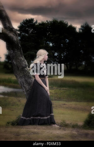 eine Frau in einem schwarzen Kleid mit Blume im Haar ist an einen Baum gelehnt. Stockfoto