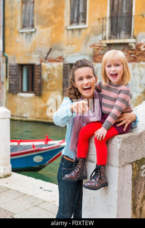 Schau, ist da Papa! Geben wir ihm ein großes Lächeln! Eine Mutter hält glücklich lachend Tochter, als sie nahe am Wasser auf einer steinernen Brücke in Venedig sitzt. Die Tochter ist stolz auf ihre rote Strumpfhose! Stockfoto