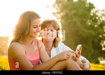 Junges Mädchen zeigt ihre Mutter Fotos auf Handy im Freien in der Natur mit Sonnenuntergang im Hintergrund