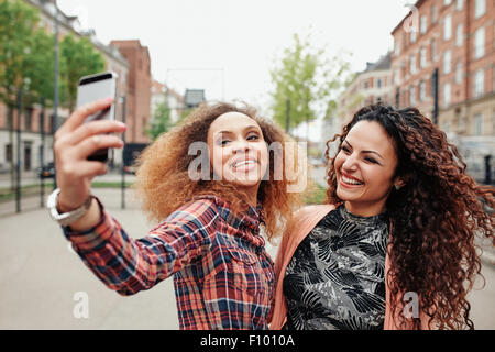 Zwei schöne junge Frauen, die ein Bild zusammen auf Stadt Straße. Junge Freundinnen nehmen ein Selbstporträt mit Handy, aus Stockfoto