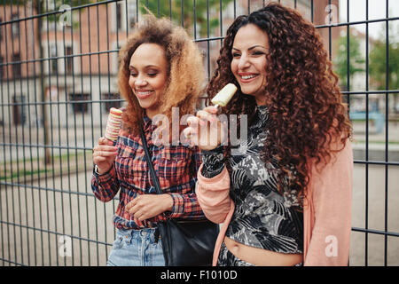 Fröhliche junge Mädchen essen Süßigkeiten Eis. Zwei junge Frauen stehen gegen einen Zaun lächelnd, im Freien. Stockfoto