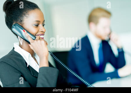 Eine schöne, schwarze, junge Frau arbeitet in einem Callcenter in einem Büro mit ihrem roten Haird Partner am anderen Ende des Schreibtisches Stockfoto