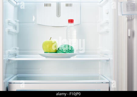 Grüner Apfel mit Maßband auf weißen Teller und eine Flasche Wasser in offenen leeren Kühlschrank. Gewicht-Verlust-Diät-Konzept. Stockfoto