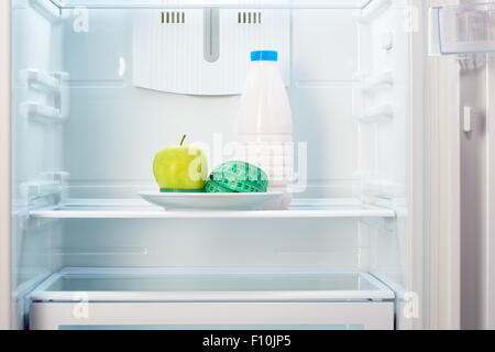 Grüner Apfel mit Maßband auf weißen Teller und eine Flasche von Joghurt im offenen leeren Kühlschrank. Gewicht-Verlust-Diät-Konzept. Stockfoto