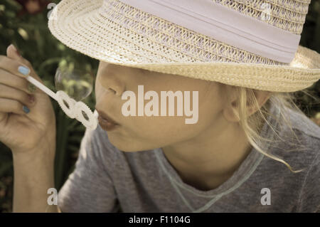 Kleines Mädchen bläst Seifenblasen einen Hut außerhalb mit Blumen im Hintergrund Stockfoto