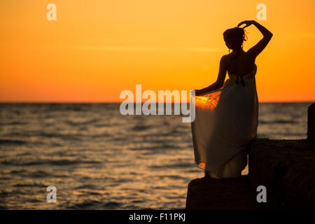 Eine Silhouette ein Frauen-Mädchen trägt einen langen fließenden Kleid posiert Modellierung als eine "griechische Göttin" von den Ozean Meer bei Sonnenuntergang Stockfoto