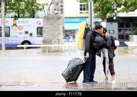 Ein paar küssen Auf Wiedersehen auf einem nassen regnerischen Nachmittag in einem geschäftigen Stadtzentrum. Die in Strömen regnen, aber der Mann hat einen Koffer und sieht zu Position aus sein Stockfoto