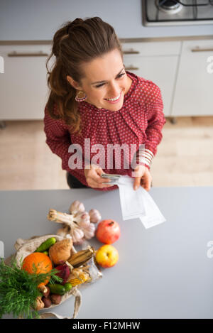 Eine elegante Frau stehen und Lächeln, Blick in die Ferne. Neben ihr auf dem Küchentisch sind frische Herbst Früchte und Gemüse. In ihren Händen hält sie Einkaufs-Listen und Bargeld. Stockfoto