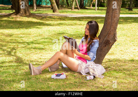 Brunette Modell tragen rosa Top und weiße Shorts sitzen auf dem Rasen im Park gelehnt auf Baum beim Lesen Buch glücklich Stockfoto