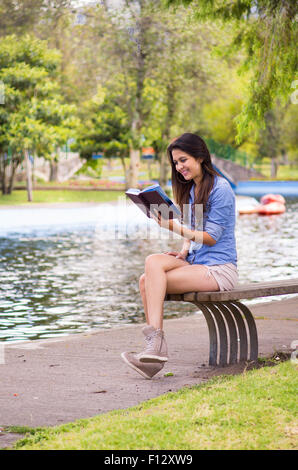 Brunette Modell tragen Jeans-Hemd und weißen Shorts in Park Umgebung entspannen, sitzen auf der Bank neben dem See Buch Stockfoto