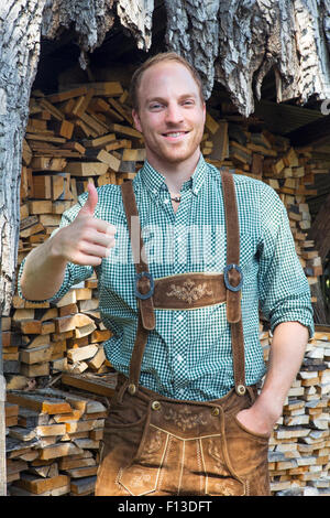 junger Mann in bayerischen Lederhosen stand vor seinen Daumen hochhalten Brennholz Stockfoto