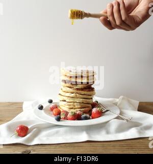 Mannes hand nieselte Honig über einen frisch zubereiteten Stapel von Pfannkuchen Stockfoto