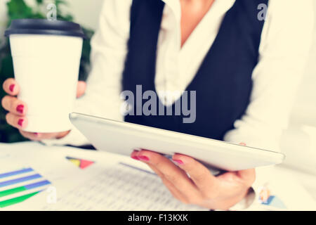 eine junge Geschäftsfrau mit einem Tablet-Computer mit einer Tasse Kaffee in der Hand, sitzen an einem Schreibtisch voller Diagramme Stockfoto
