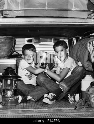 1960ER JAHRE ZWEI LÄCHELNDEN JUNGEN BRÜDER UMARMEN HAUSTIER HUND BLICK IN DIE KAMERA SITZT AUF DER RÜCKSEITE GEPACKTEN KOMBI AUTO BEREIT FÜR CAMPING Stockfoto