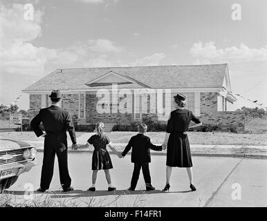 1960ER JAHREN VIERKÖPFIGE FAMILIE GESEHEN VON HINTEN STAND VOR NEUEN VORSTADTHAUS HAND IN HAND Stockfoto