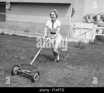 1950ER JAHREN LÄCHELNDEN BLONDEN FRAU IN SHORTS MÄHEN RASEN MIT PUSH-MÄHER Stockfoto