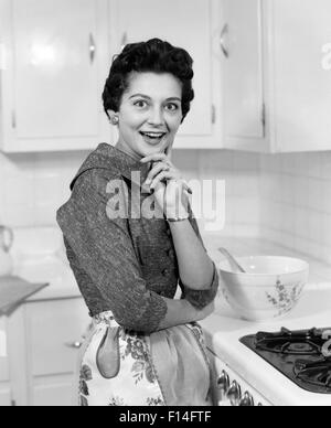 1950ER JAHREN LÄCHELND BRÜNETTE FRAU HAUSFRAU NEBEN HERD TRÄGT SCHÜRZE IN KÜCHE ÜBERRASCHT, MIMIK, BLICK IN DIE KAMERA Stockfoto