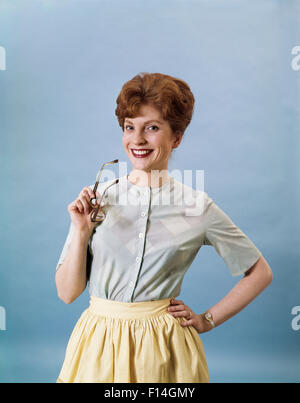 1960ER JAHREN LÄCHELNDE FRAU TRÄGT SCHÜRZE BLICK IN DIE KAMERA GESTIKULIEREN MIT BRILLE Stockfoto