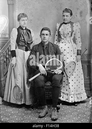 1890ER JAHRE PORTRAIT ZWEI FRAUEN EIN MANN POSIERT MIT TENNISSCHLÄGER Stockfoto