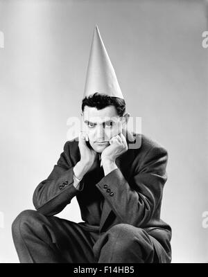 1950ER JAHRE DISGUNTLED MANN TRÄGT DUNCE CAP BLICK IN DIE KAMERA Stockfoto