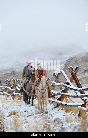 Cowboy und Cowgirl Reiten auf Pferden neben Zaun in Schnee, Rocky Mountains, Wyoming, USA Stockfoto