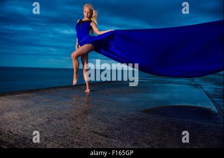 Open Air Location Fotografie - ein groß schlank blond blonde Frau Mädchen Model posiert im Freien an einem nassen Abend mit einem langen blauen fließenden Kleid schweben im wind Stockfoto