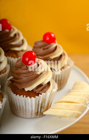 Bananen-Muffins mit Schokolade/Vanille wirbeln Frosting und glasierten Kirsche obenauf Stockfoto