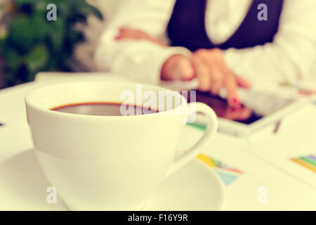 Nahaufnahme von einer Tasse Kaffee auf dem Schreibtisch voller Diagramme und eine junge Geschäftsfrau mit einem Tabletcomputer Stockfoto