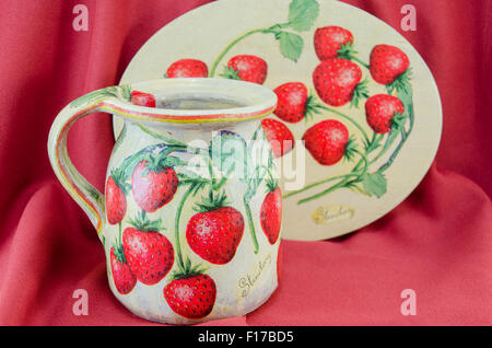 Serviettentechnik dekoriert Erdbeer Muster Ton Krug und Teller auf rotem Stoff Hintergrund Stockfoto