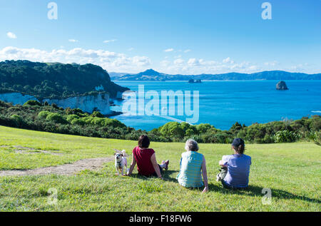 3 Personen und ihr Hund entspannt auf dem Rasen und Blick auf Edelstein-Bucht und die Coromandel-Halbinsel Stockfoto