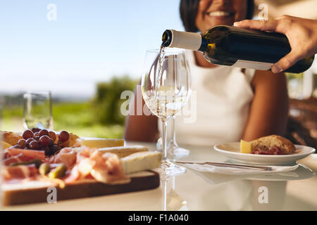 Hände eines Mannes mit einer Frau im Hintergrund am Weingut lächelnd zwei Gläser aus Flasche Weißwein herein. Fokus auf Gläser und Stockfoto