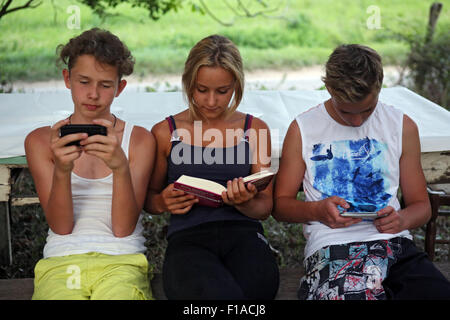 Lago di Bolsena, Italien, spielen die Jungs mit ihren Smartphones während eines Mädchens in einem Buch lesen Stockfoto